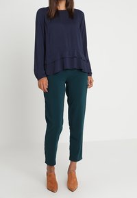 Tummansininen pitkähihaainen paita, jossa on röyhelöhelma, yhdistetty mehukkaan teal-värisiin slim fit -housuihin ja vaaleanruskeisiin korkokenkiin.