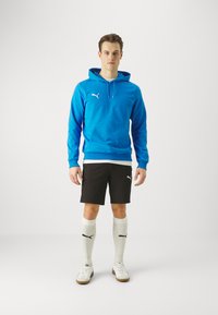 Kék kapucnis pulóver kenguruzsebbe, fekete rövidnadrág, fehér sportcipő, és világos színű focizokni logóval. Egyszerű dizájn kontrasztos színekkel.
