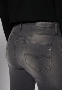 Jeans em denim cinza com um acabamento desbotado, apresentando uma etiqueta com a inscrição "Replay Hyperflex" e costuras subtis nos bolsos traseiros.