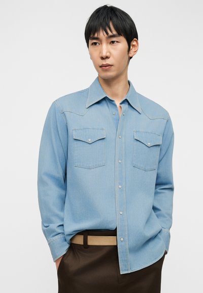 Ανοιχτά γαλάζιο πουκάμισο από τζιν με μυτερό γιακά, δύο τσέπες στο στήθος και ορατή λωρίδα με κουμπιά; συνδυασμένο με καφέ παντελόνια και μπεζ ζώνη.