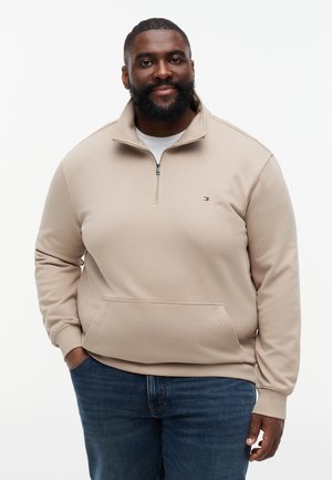 Muži s bradou, oblečený v béžovej mikine so zipsom po štvrťku a modrých džínsoch, stojaci s jednou rukou v vrecku na bielom pozadí.