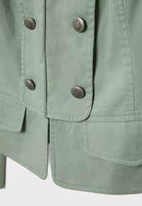 Veste vert clair vue de face montrant des boutons argentés décoratifs en deux rangées verticales et des surpiqûres détaillées sur les poches et l'ourlet.