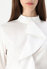 Hvit bluse med høy krage og store, brettede volanger foran. Glatt stoff med en svakt glinsende overflate.