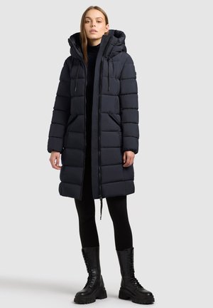 Marineblauer Puffercoat mit schulterlangem Schnitt, ausgestattet mit einer Kapuze, frontalem Reißverschluss und Seitentaschen; kombiniert mit schwarzen Leggings und klobigen schwarzen Stiefeln.