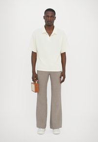 Krämfärgad kortärmad polo, ljus texturerad tyg, i kombination med högmidjade, vida beige rutiga byxor. Håller en liten brun handväska.