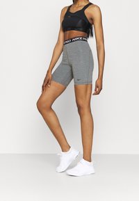 Szare spodenki sportowe z wysokim stanem i czarnym logo Nike, w połączeniu z czarnym biustonoszem sportowym i białymi trampkami, tworząc strój sportowy.