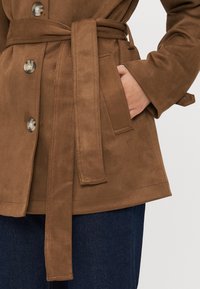Braune Wildlederjacke mit großen Metallknöpfen, einem gebundenen Gürtel in der Taille und Seitentaschen, die eine weiche Textur und ein strukturiertes Design aufweist.