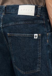 Jean en denim bleu foncé avec des coutures orange, un patch blanc avec un texte japonais sur la ceinture, et une petite étiquette logo coléoptère sur la poche arrière.
