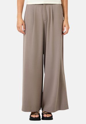 Personne portant un pantalon taupe ample à jambes larges associé à des sandales noires à brides, debout devant un fond clair uni.