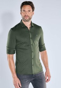 Olivgrünes Hemd zum Zuschließen mit langen Ärmeln, die am Bündchen hochgekrempelt sind, mit einem dezenten Logo, aus weichem Stoff gefertigt und kombiniert mit grauer Hose.