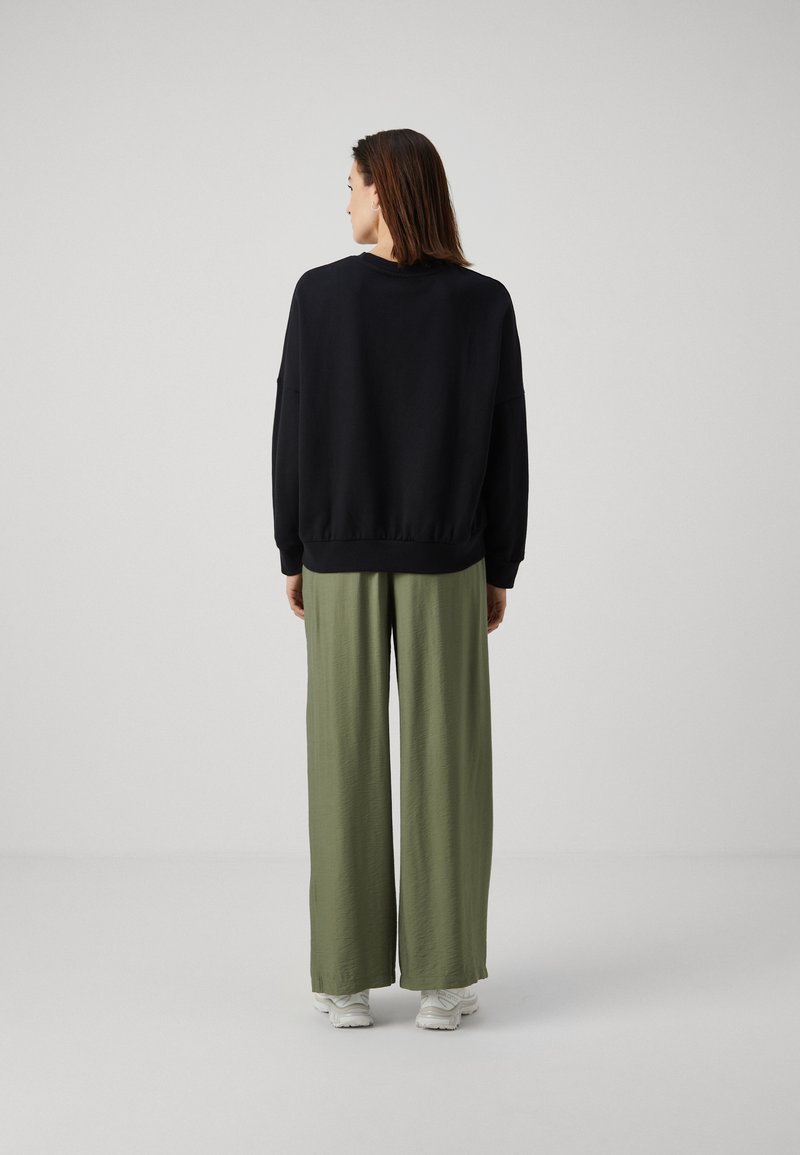 Felpa nera con polsini e orlo a coste, abbinata a pantaloni ampi verde oliva. Tessuto morbido, vestibilità rilassata. Come calzature, scarpe da ginnastica bianche.