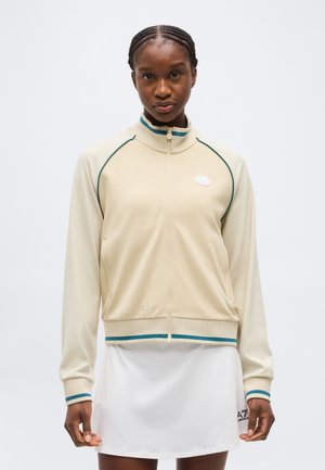 Punutud juustega naine, kes kannab beeži lukuga jope tealsete äärtega ja valget seelikut, seisab ühetaolise heleda tausta ees.