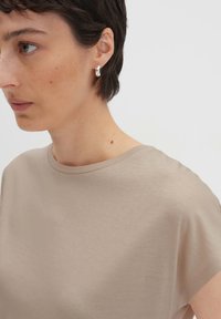 Top beige a maniche corte realizzato in tessuto morbido con scollatura rotonda. Presenta una vestibilità rilassata e una texture liscia. Orecchino a cerchio in argento visibile.
