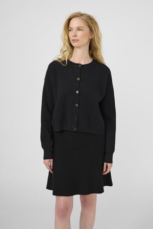 Plava žena u crnom kardiganu na dugmad i odgovarajućoj crnoj suknji, stoji naslonjena na jednostavnu svijetlosivu pozadinu.