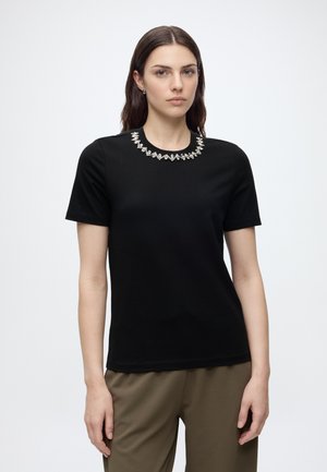 Femme aux longs cheveux foncés portant un haut noir à manches courtes avec un col bijouté et un pantalon vert olive sur un fond clair uni.