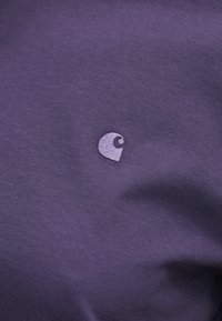 Violets kokvilnas audums ar mazām izšūtām emblēmām gaišākā tonī, demonstrējot vienkāršu, gludu faktūru un minimālisma dizainu.