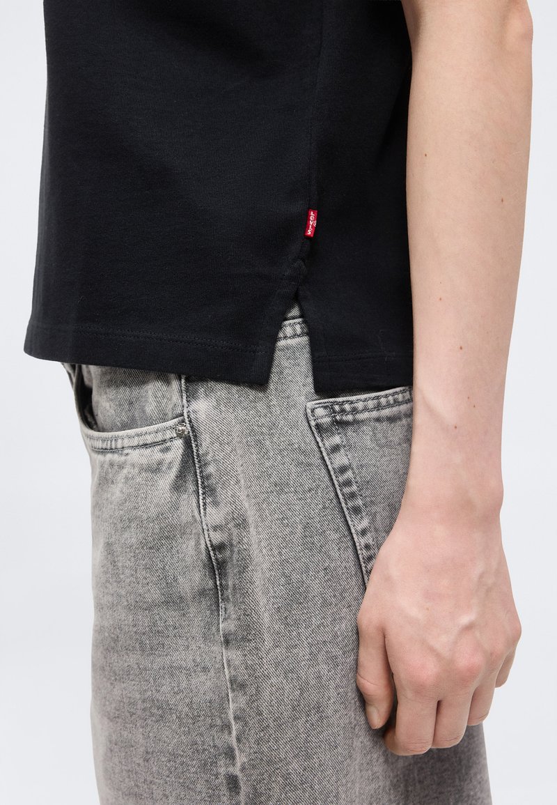 Personne portant un t-shirt noir avec une fente latérale et une petite étiquette rouge, associé à un jean gris clair délavé, le bras reposant le long du corps.