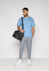 Ανοιχτογάλανο κοντομάνικο μπλουζάκι, γκρι στενά φαρδιά παντελόνια φόρμας, λευκά αθλητικά παπούτσια και μαύρο σάκο ταξιδίου με ιμάντα ώμου.