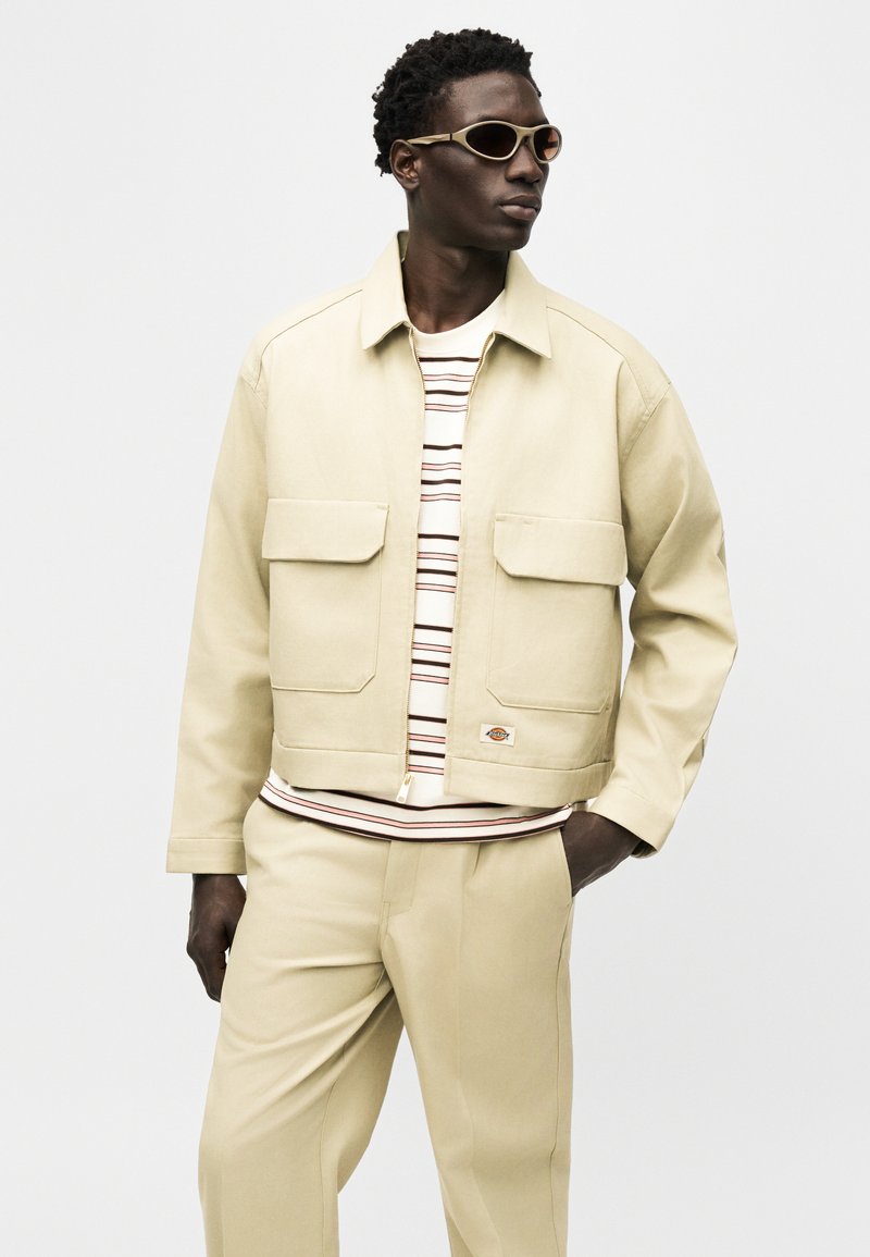 Homme portant une veste et un pantalon beige avec une chemise à rayures et des lunettes de soleil ovales, debout avec une main dans la poche, regardant vers la droite.