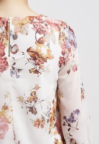 Blusa floreale realizzata in tessuto leggero e trasparente con colori pastello. Il design include una schiena a chiave con una cerniera metallica visibile e maniche lunghe.