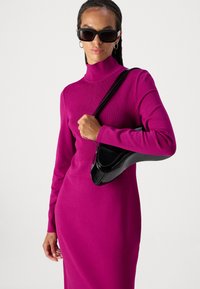 Fuchsia-Kleid mit hohem Kragen und langen Ärmeln, das eine figurbetonte Passform hat. Das Model hält eine schwarze, glänzende Handtasche mit geschwungener Form.