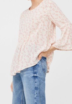 Femme portant un chemisier rose clair à manches longues avec un motif floral et un jean bleu, posant avec une main dans la poche.