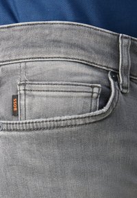 Los jeans de mezclilla gris presentan un diseño de bolsillo estándar, con costuras visibles y una pequeña etiqueta naranja "BOSS" en el bolsillo trasero.