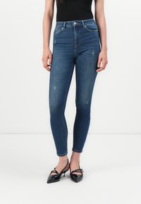Höga blå jeans i denim, med smal passform, lätt blekning och fem-ficksdesign; tillsammans med svarta patenterade slingback-skor.