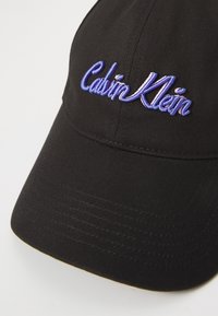 Căciulă de baseball neagră din bumbac, cu vizor curbat. Prezintă un logo brodat "Calvin Klein" în mov și roz pe față.