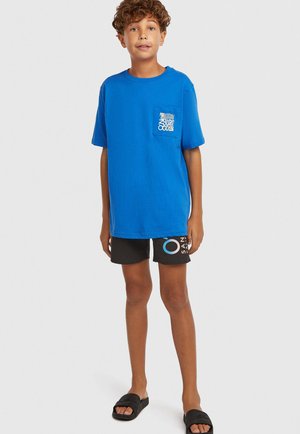 Blå bomuld t-shirt med en lomme med hvid tekst, kombineret med sorte shorts med et grafisk design og sorte slidesandaler.