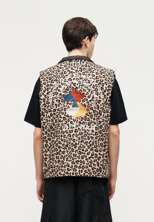 Personne portant un gilet sans manches à imprimé léopard avec des patchs colorés brodés et du texte au dos, par-dessus une chemise noire.