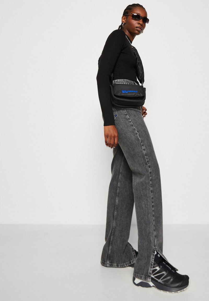 Pull à mailles noir, jean taille haute gris, sac bandoulière noir avec logo bleu, et baskets noires. Des lunettes de soleil complètent le look moderne.