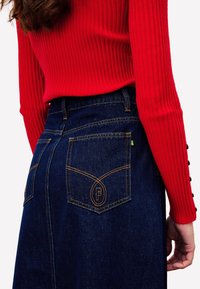 Jupe en denim bleu foncé avec des coutures jaune-orange, dotée de deux poches arrière avec un logo de marque, associée à un pull rouge à côtes.