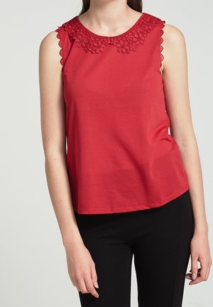 Mouwloos rood topje met bloemige kanten afwerking rond de halslijn en schouders, gedragen met zwarte nauwsluitende broek, getoond van nek tot heupen.