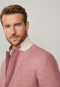 Chaqueta de lino rosa con cuello alto y detalles en crema, que presenta un tejido texturizado y un bolsillo lateral. Modelo con una camisa de color azul claro debajo.
