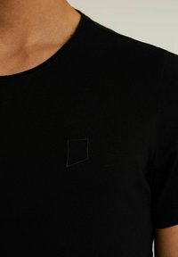 Černé tričko s krátkým rukávem vyrobené z žebrované tkaniny, na levém prsu je malá, vyšitá čtvercová logo. Hladká textura.