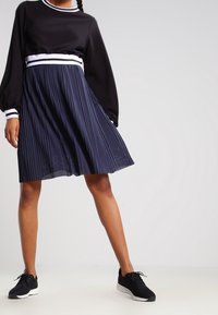 Svart långärmad topp med vita ränder vid halsringningen, parat med en marinblå plisserad kjol. Modellen bär svarta träningsskor.