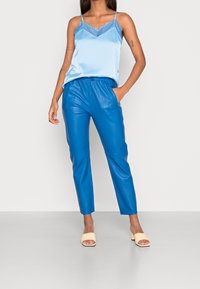 Light blue satin camisole with lace trim, paired with high-waisted, shiny blue faux leather trousers and beige block-heeled sandals.