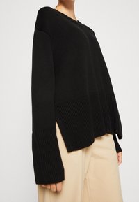 Svart stickad tröja med lös passform, med ribbad textur och vida ärmar. Nederkanten har sidoflikar för ökad rörelsefrihet och komfort.