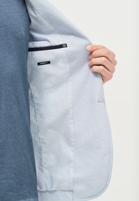 Světle modrý blazer s texturovaným materiálem, zapínání na jeden knoflík a vnitřní kapsa označená "MODERNÍ STŘIH." Odkazováno částečně otevřený.