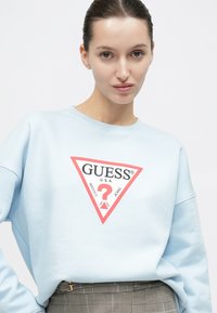 Νέα γυναίκα με μαλλιά δεμένα πίσω φοράει γαλάζιο φούτερ με το λογότυπο Guess περιμετρικά κόκκινο και γκρι καρό παντελόνι, σε λευκό φόντο.