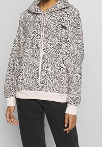 Person som bär en ljusrosa huvtröja med abstrakta svarta snirklar och en framficka, tillsammans med mörka byxor.