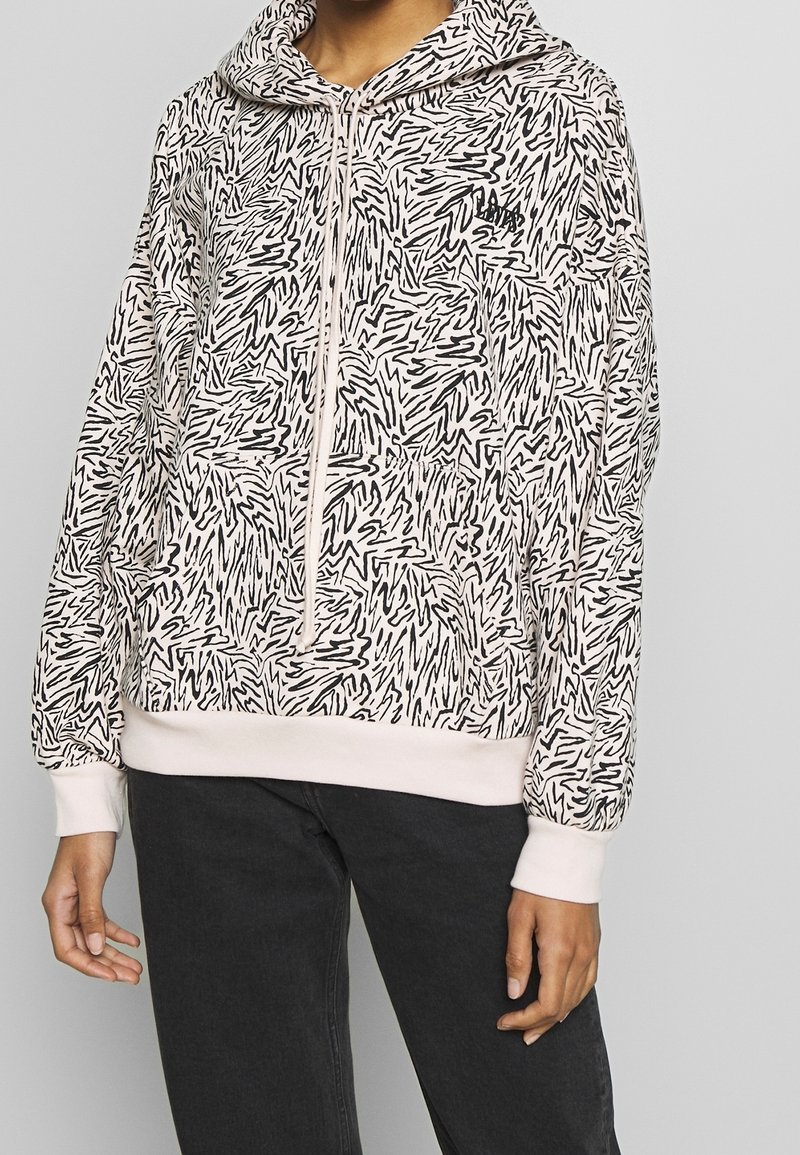 Person som bär en ljusrosa huvtröja med abstrakta svarta snirklar och en framficka, tillsammans med mörka byxor.