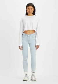 Haut blanc cropped à manches longues avec détail de texte, associé à un jean skinny bleu clair. Le modèle porte des baskets blanches avec des accents verts.