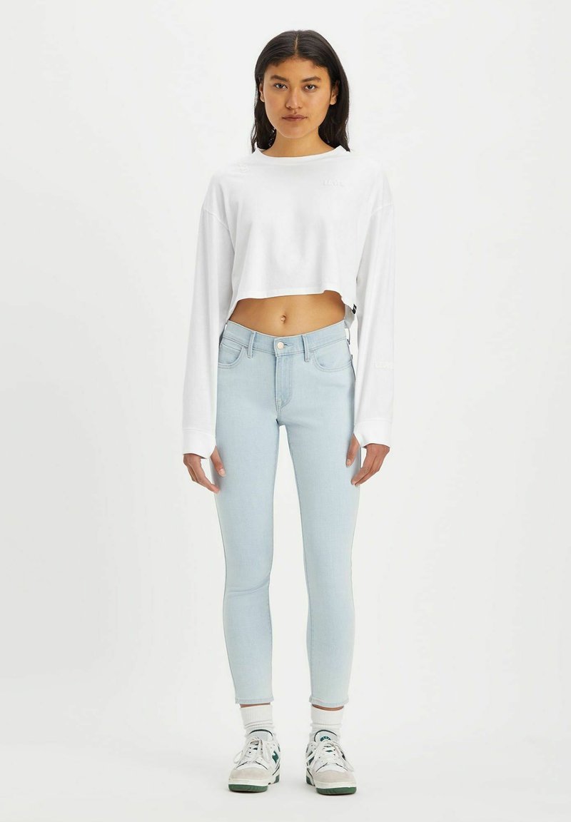 Haut blanc cropped à manches longues avec détail de texte, associé à un jean skinny bleu clair. Le modèle porte des baskets blanches avec des accents verts.