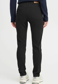 Black jeans with a slim fit, featuring a smooth texture, two back pockets, and a tan label at the waistband. Paired with white sneakers.