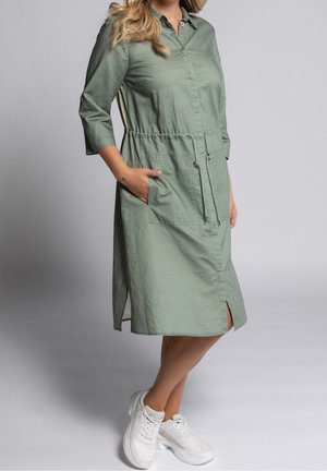 Frau trägt ein hellgrünes Midikleid mit Fronttaschen und Kordelzug an der Taille, kombiniert mit weißen Sneakers vor einem einfarbigen Hintergrund.