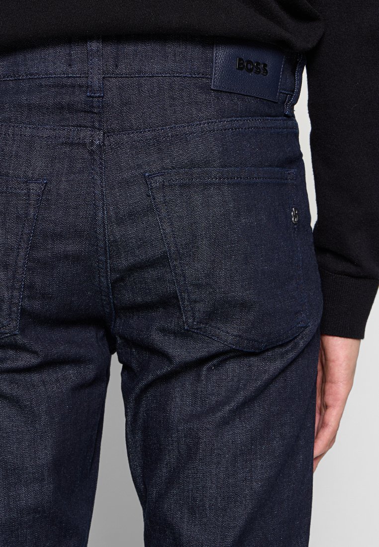 Dunkelblaue Jeans mit schlanker Passform, ausgestattet mit Gesäßtaschen und einem schwarzen Lederlogo-Tag. Glatte Textur und klassische blaue Farbe.