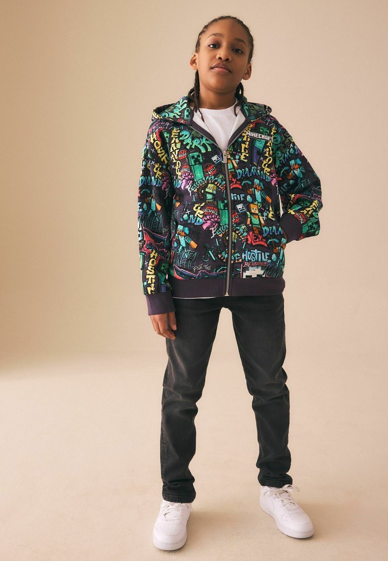 Jonge persoon staat met één hand in de zak, draagt een kleurrijke Minecraft-thema zip-up hoodie, zwarte spijkerbroek en witte sneakers, neutrale achtergrond.