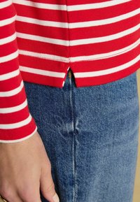 Czerwono-biała bluzka z długim rękawem w paski z małym rozcięciem z tyłu, noszona z niebieskimi dżinsami. Widoczna ręka oparta na dżinsach.